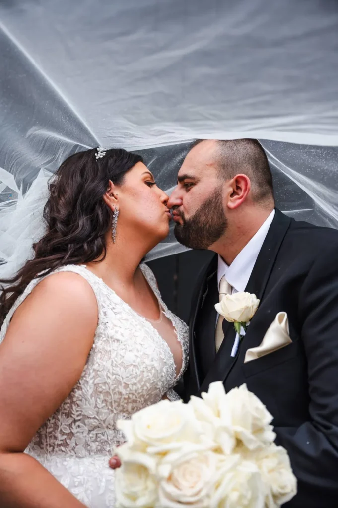 Bride and groom kiss under the veil holding white roses