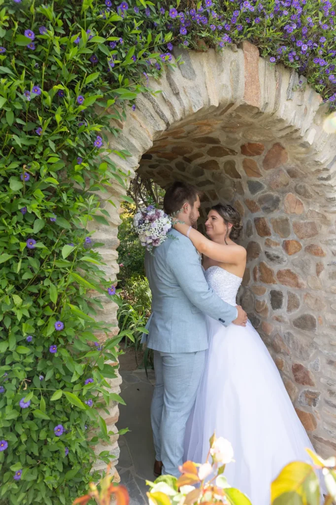 Bride and groom embracing under a flower-covered stone archway