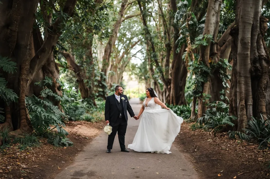 Bride and groom walking hand in hand down a tree-lined avenue